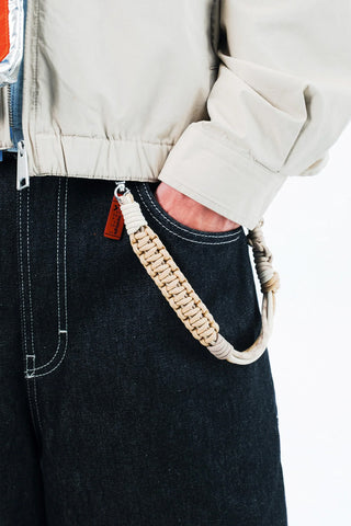Close-up of a person's jeans with a taupe and cream braided rope wrist strap clipped on their jeans on a white background