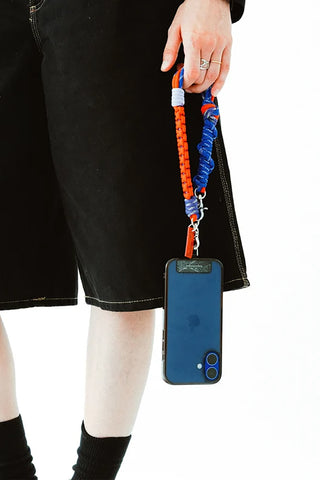 A close-up of a person wearing black denim shorts holding a blue smartphone with an orange and blue braided phone strap attached.