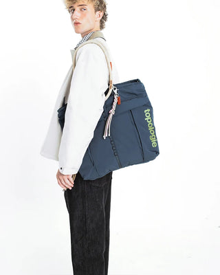A model wearing a white jacket and black jeans carrying a navy tote bag with a woven rope keychain on a white background.