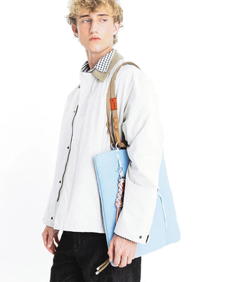 A person wearing a white jacket and black pants, carrying a light blue tote bag with a rope keychain on a white background.