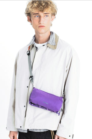 A model wearing a light beige jacket carrying a purple crossbody pouch bag.