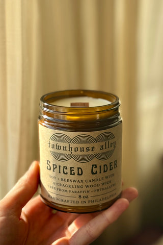 Candle jar labeled 'Spiced Cider' held by a hand against a neutral background