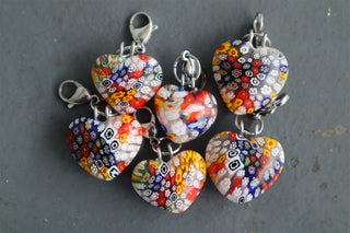 Five heart-shaped glass charms with silver clasps, featuring vibrant multicolor Millefiori patterns, on a dark gray textured surface.
