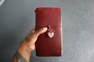Hand holding leather journal with millefiori patterned heart shaped pendant against a gray textured surface