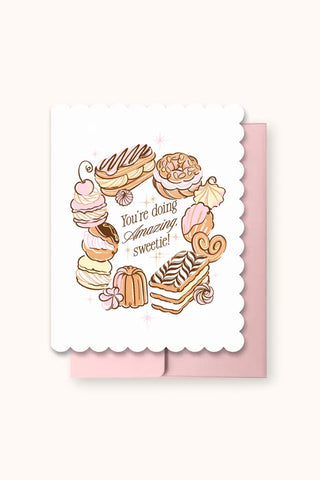 Greeting card featuring an illustration of assorted pastries and phrase 'You're doing Amazing sweetie!', shown with a light pink envelope behind it
