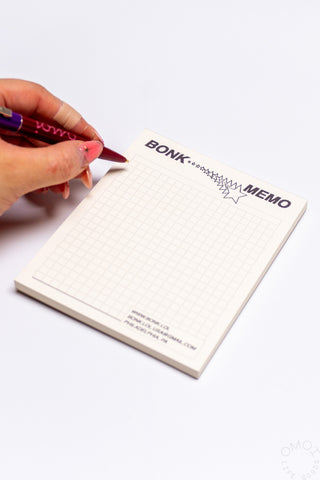 Hand holding a pen over a small notepad with 'BONK' branding on a white background