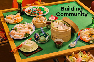 Table with various Chinese food items with mahjong tiles in between and 'Building Community' text on a green mat.
