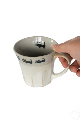 Hand holding a beige ceramic mug with cat parade design on a white background