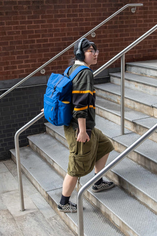 Person with a blue backpack standing on a set of stairs against a brick wall.