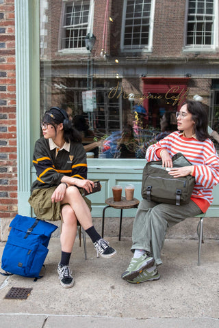 Two people sitting on a sidewalk with backpacks and coffee cups, in front of a shop window.