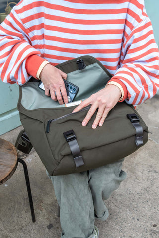 Person holding a green bag with a phone inside, wearing a striped shirt and light pants.