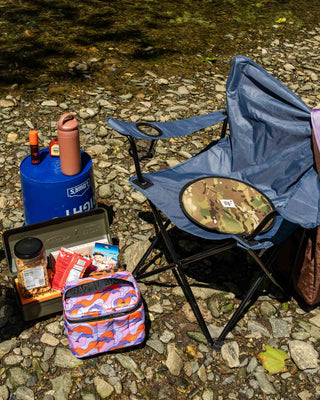 Peace Cabin ECLIPSE Cordura Seat Pad Woodland resting on a blue folding chair by a rocky creek with outdoor snacks and a water bottle nearby.