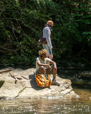 people standing on rock near lake surrounded by trees, one standing with hat and bag, other person sitting with bag
