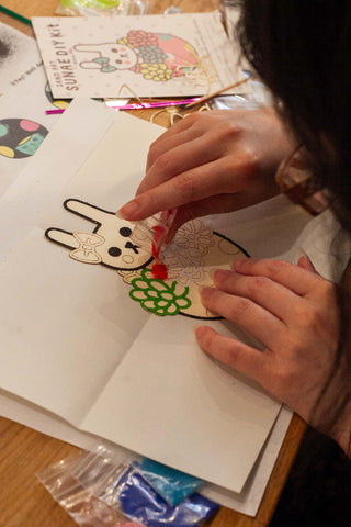 A person carefully pours colorful sand onto a Naoshi sunae sand art kit. Their design is one of a bunny in a flower pot.