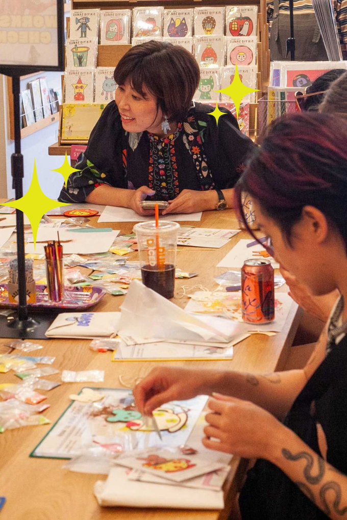 People drawing at a craft table with pens, papers, and paint jars