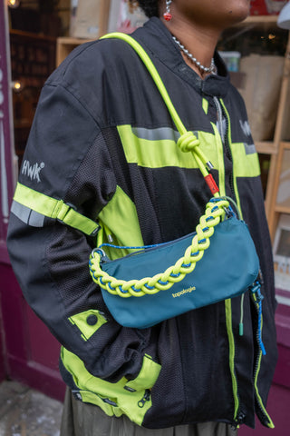 Person wearing a black and green jacket with a blue bag featuring braided design.